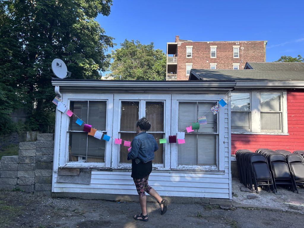 a person wearing a jean jacket, a skirt and floral leggings reads colorful pieces of paper hung on a string, on a white building with crumbling paint. 